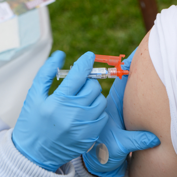 Healthcare worker in blue gloves injecting flu vaccine into a patient's upper arm with syringe and orange safety cap