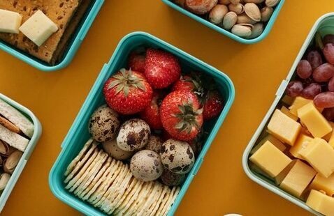 Teal lunch boxes on yellow background with sandwiches, cheese cubes, grapes, strawberries, crackers, nuts and quail eggs