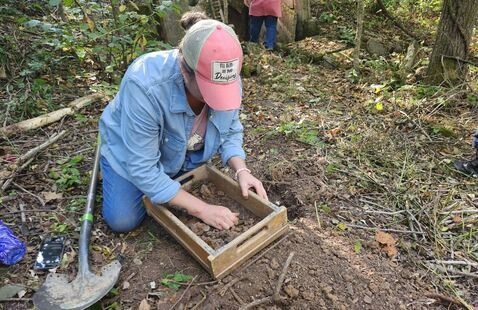 Archaeologist kneeling in woods, sifting soil through a wooden screen beside a shovel and dirt pile.