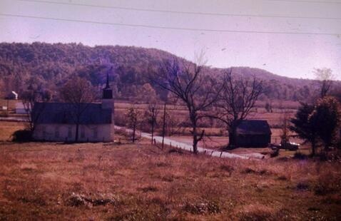 Small white church with steeple beside country road and barn, leafless trees and grassy field with wooded hills behind. A schoolhouse sits across the gravel road from the church.
