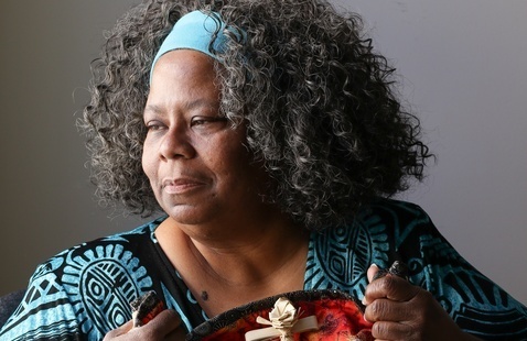 Cookie Washington, a woman with gray curly hair and blue headband, holds a colorful quilt with a small woven cross.