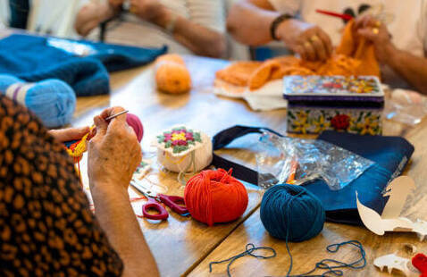 Hands of knitters at a table knitting with orange yarn, red and blue yarn balls, pincushion, scissors, and a decorative tin.