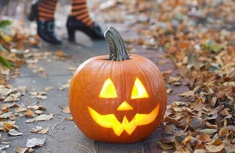 Lit pumpkin on sidewalk in front of legs of person dressed in witch costume