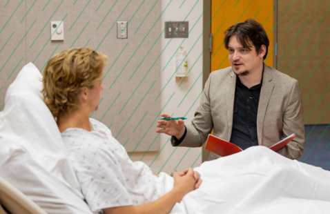 A student sits next to a patient in a hospital setting