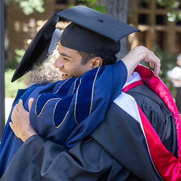Stanford Master of Liberal Arts graduate in cap and gown hugs faculty in blue academic robe at graduation.