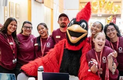 Seven student staff in maroon shirts pose indoors with a red cardinal mascot at a table; one staff member displays keychains.