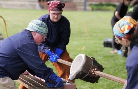 Three people in bandanas and gloves pouring molten metal from a crucible into a mold on a grassy lawn