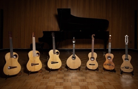 Seven acoustic and other stringed instruments on stands arranged on parquet floor before a grand piano and wood-paneled wall