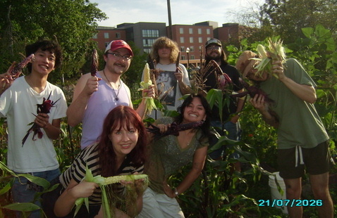 Seven people holding harvested corn in a community garden with Kalapuya Ilihi dorm building in the background, 21/07/2026