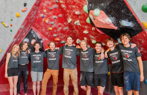 Nine climbers wearing 'WESTERN COLORADO' shirts pose in front of red indoor bouldering wall.