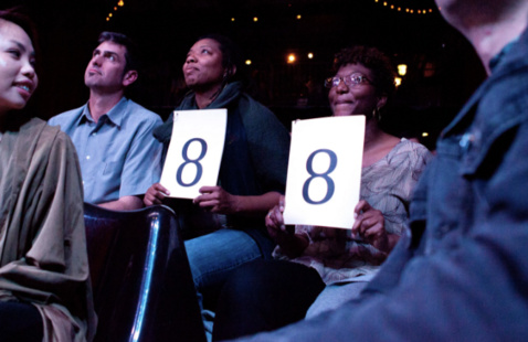 audience members in a crowd hold up papers reading "8"