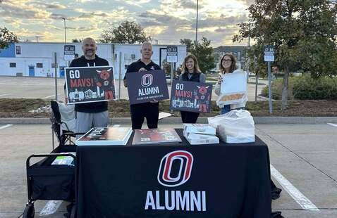 Four people behind a table with ALUMNI tablecloth holding signs 'GO MAVS!' and 'ALUMNI FOREVER MAVERICKS' in a parking lot