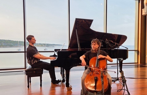 A cellist seated playing cello beside pianist at grand piano by large windows overlooking a bay with sailboats.