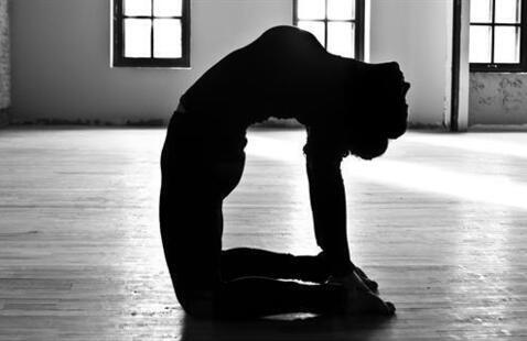 Silhouette of person in camel yoga backbend on wooden studio floor with tall windows and sunlight