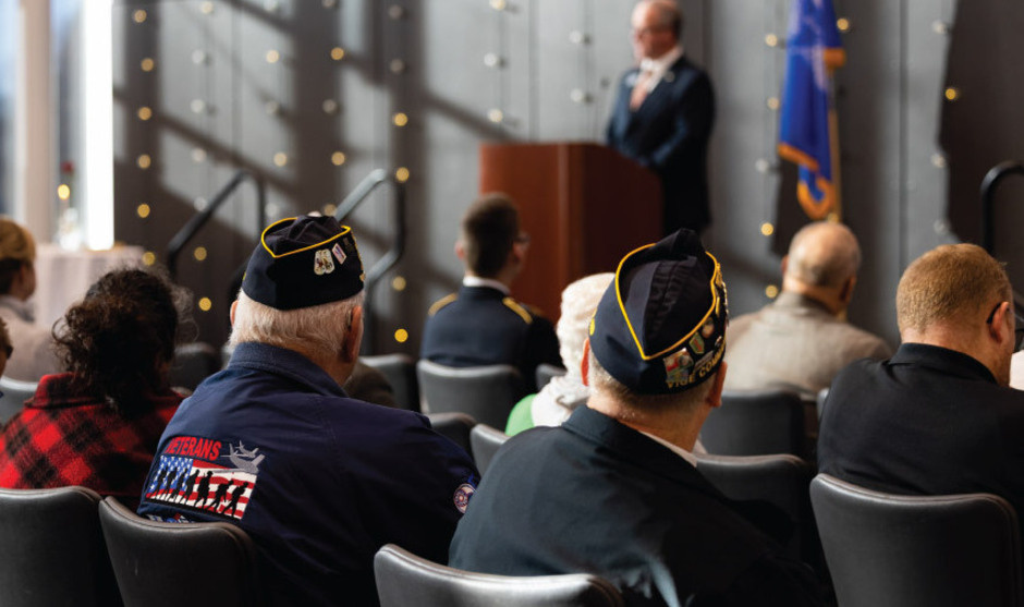 People seated watching speech on stage, veterans wearing garrison caps